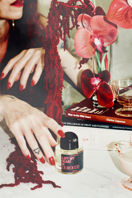 A perfume bottle labeled 'LEFT MY HEART IN MOROCCO' sits on a white surface, surrounded by red yarn, pink orchids, stacked books, and a glass of amber liquid. A person's hand with red nail polish reaches toward the bottle.