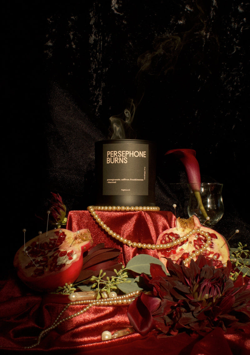 A matte black glass candle with a label reading 'PERSEPHONE BURNS' placed on a table with red fabric, surrounded by decorative elements and a glass of wine in the background.